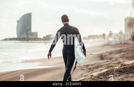 adult man in wetsuit carrying surfboard walking on sandy beach in sunlight, Barcelona Stock Photo
