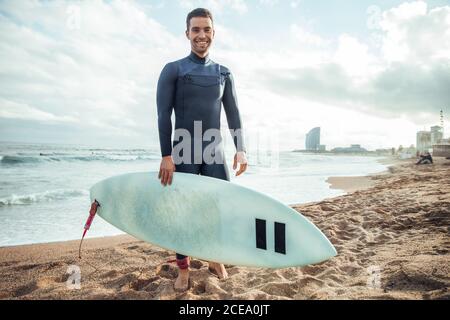 man wearing wetsuit and standing with surfing board on sandy beach, Spain Stock Photo