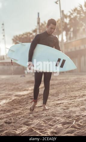 Happy adult man in wetsuit carrying surfboard walking on sandy beach in sunlight, Barcelona Stock Photo