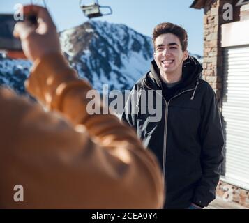 unrecognizable person using smartphone to take picture of smiling young man on sunny day on ski resort Stock Photo