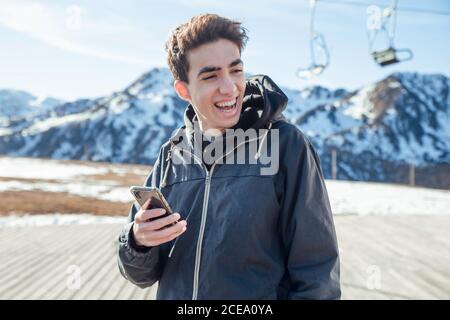 Handsome young man with smartphone laughing and looking away while standing on background of snowy mountains on ski resort Stock Photo