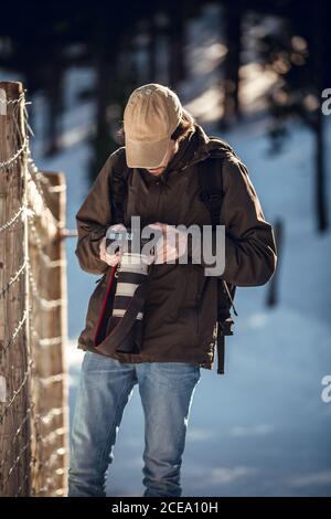 Young man in sunglasses and cap with backpack looking at professional camera screen between winter forest in Cerdanya, France Stock Photo