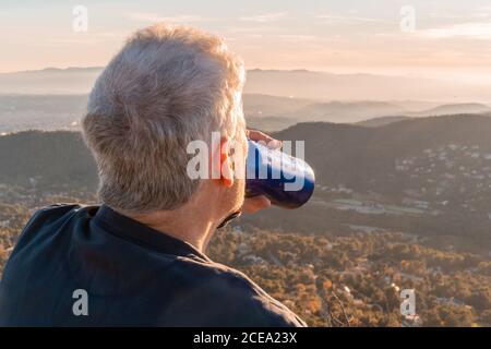 Back view of senior traveler drinking fresh beverage from bottle while admiring view of majestic countryside during sundown Stock Photo