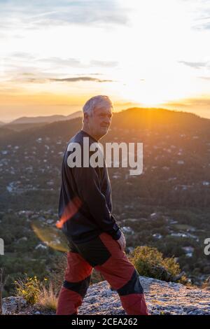 Side view of elderly man in casual outfit standing in a rocky cliff and admiring view of majestic mountains and beautiful evening sky Stock Photo