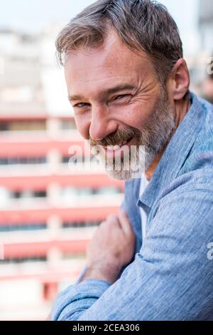 Portrait of bearded man with wrinkles smiling and looking at camera on blurred building background Stock Photo