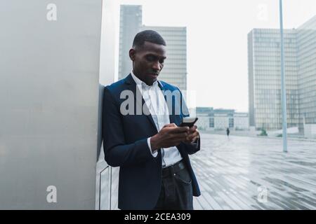 Handsome young African-American male in elegant suit standing relaxed leaning on wall of building and concentrating on working with mobile phone with city of glass and concrete after rain on background Stock Photo