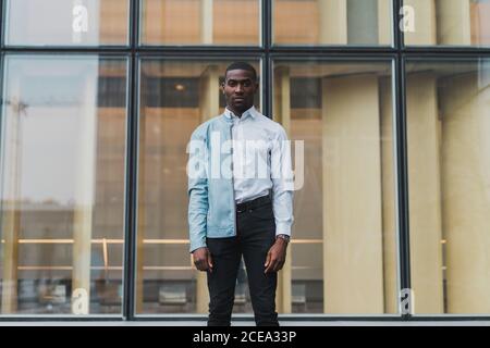 Handsome young African-American man in elegant clothes and blue leather jacket on one shoulder standing relaxed at glass wall of building and looking at camera Stock Photo