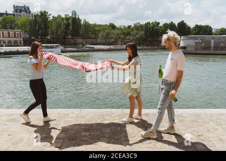 Side view of two young women in casual clothes spreading blanket together on waterfront while stylish man looking at them holding bottles of water in hands Stock Photo