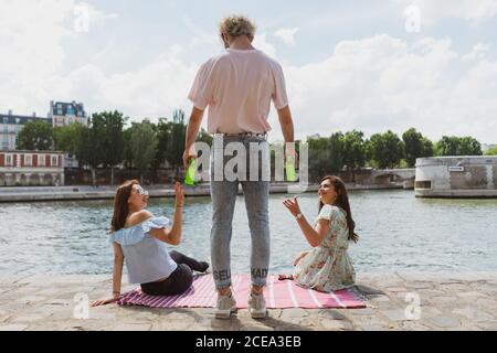 Back view of stylish man standing giving bottles of water to young women in casual clothes sitting on waterfront Stock Photo