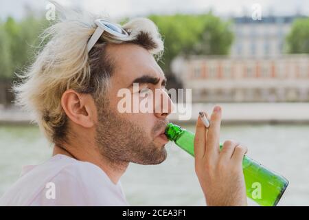 Side view of�blond bearded male holding cigarette in hand while drinking beer on street on blurred background Stock Photo