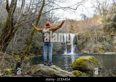 Back view of man with backpack stretching out arms and admiring beautiful waterfall while standing on boulder near forest lake in Spain Stock Photo