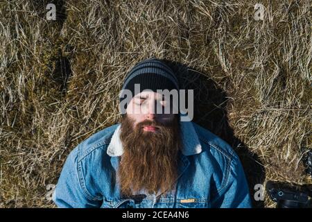 View from above of bearded young male in hat with close eyes lying on grass in Iceland Stock Photo