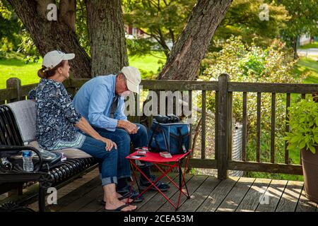 Chesapeake City, MD, USA 08/25/2020: An elderly caucasian couple wearing baseball hats shirts and jeans is sitting on pillows on a metal park bench in Stock Photo