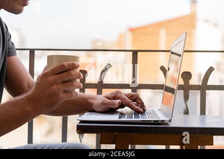Crop man with cup of coffee and using laptop on balcony Stock Photo