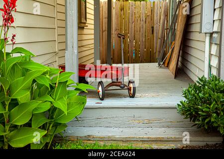 A red old rusty toy wagon was left on the doorway of an old house. The wooden porch the toy was left has grey paint which shows signs of aging. There Stock Photo