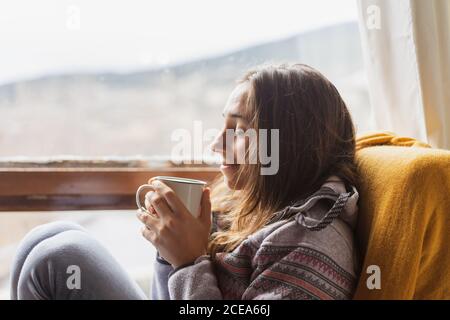 Side view of cute female in warm jumper smiling and enjoying hot beverage while sitting in a armchair near huge window at home Stock Photo