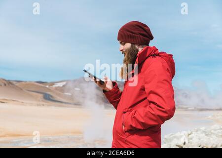 Side view of bearded male in warm bright clothes using mobile phone standing in front of steaming hot spring in Namaskardh in Iceland Stock Photo