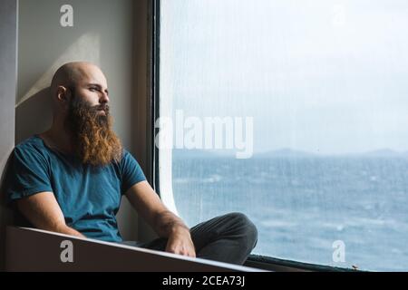 Thoughtful adult bearded man sitting at the window and looking away. Stock Photo