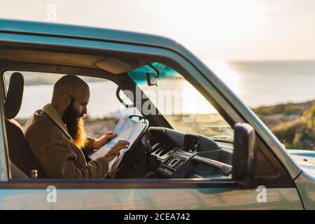 Side view of adult bearded man resting in the car and reading map at the seaside. Stock Photo
