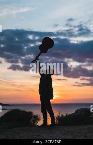 Side view of bearded man having fun and posing with guitar in sunset lights on ocean coast. Stock Photo