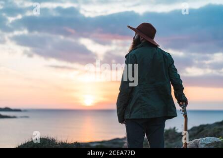 Back view of bearded tourist man standing and leaning on guitar on coast at the ocean in sunset. Stock Photo