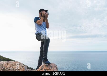Cheerful bearded man in hat sitting on stone and taking selfie at seaside. Stock Photo