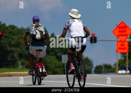 A senior couple wearing casual shorts and t shirts as well as hats is riding bikes near a highway on a sunny summer day. Man has a foldable pedal bike Stock Photo