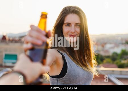 Crop view of hand giving bottle of cool beer to beautiful young female standing on rooftop and looking at camera on blurred backlit background Stock Photo