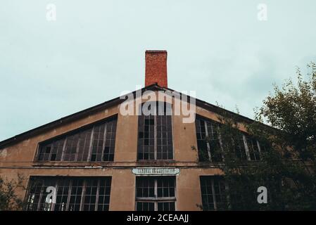 From below red facade of aged broken construction with big windows and chimney near trees and cloudy heaven Stock Photo