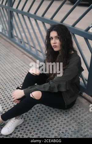 Pretty young Woman with curly hair looking at camera while sitting on weathered metal path on city street Stock Photo