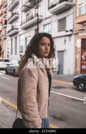 Beautiful young Woman with curly hair looking at camera while standing near modern building on city street Stock Photo