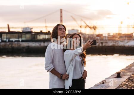 Guy near attractive lady with mobile phone and taking selfie near water surface in sunny day Stock Photo