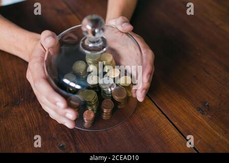 From above shot of crop hands covering stacks of coins with glass lid on wooden table Stock Photo