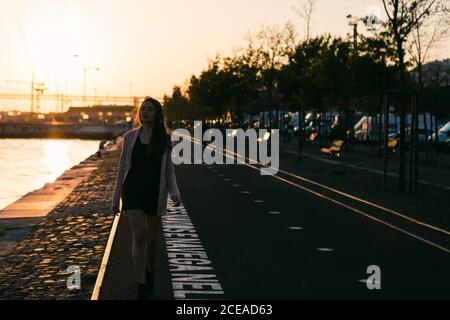 Attractive charming lady going on promenade near water and alley with benches at sunset Stock Photo