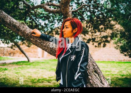 Pretty Woman in black leather jacket and with red hair standing under tree in park looking confidently away Stock Photo