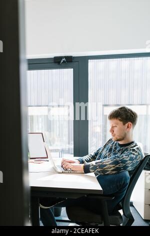Side view of male in casual clothes sitting comfortably on office chair and typing on laptop keyboard on white desk with large window on background shot made through open door Stock Photo