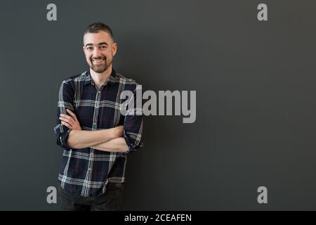 Handsome middle-aged bearded male with grey hair on temples in dark casual checkered shirt standing with crossed arms smiling and looking at camera over black background Stock Photo
