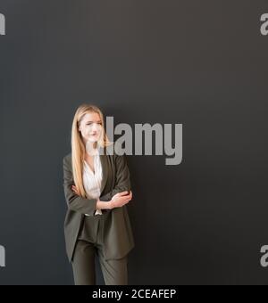 Beautiful blond young Woman in dark elegant suit standing confidently with crossed arms and looking at camera smiling on black background Stock Photo