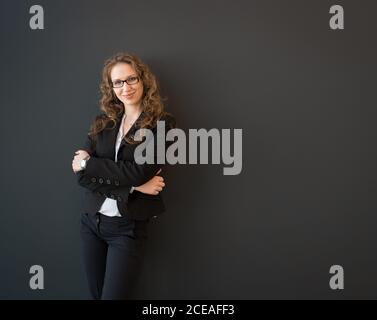Beautiful blond young Woman in dark elegant suit standing confidently with crossed arms and looking at camera smiling on black background Stock Photo