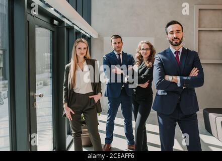 Group of office colleagues in elegant dark suits standing together relaxed with crossed arms and looking at camera smiling in spacious room at glass wall and door Stock Photo