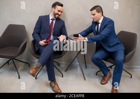 Two elegant men wearing suits and sitting on chairs in office sharing smartphone and having fun in leisure Stock Photo
