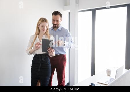 Stylish smiling man and Woman standing in light office and using tablet together while having discussion Stock Photo