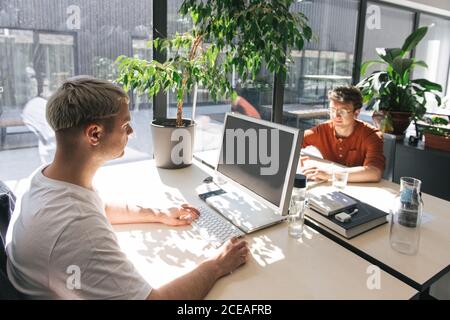 Men sitting at tables in front of each other inside of open space office and working on computers in sunlight Stock Photo