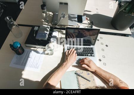 Crop shot from above of man sitting at table and working on laptop in bright sunlight inside of office Stock Photo