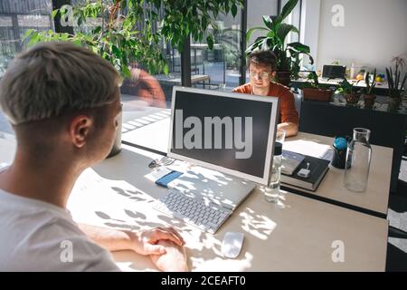 Men sitting at tables in front of each other inside of open space office and working on computers in sunlight Stock Photo