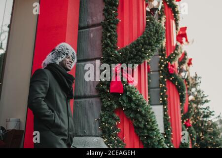From below shot of excited African American male in warm clothes while standing on city street near building and conifer tree decorated for Christmas Stock Photo