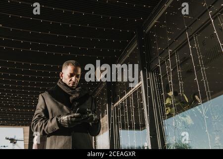 From below shot of handsome African American male using modern smartphone while standing near modern building decorating for Christmas with fairy light garlands Stock Photo