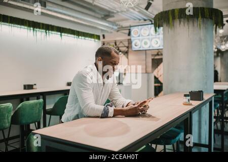 Side view of handsome African American guy smiling and using modern smartphone while sitting at table in stylish cafe Stock Photo