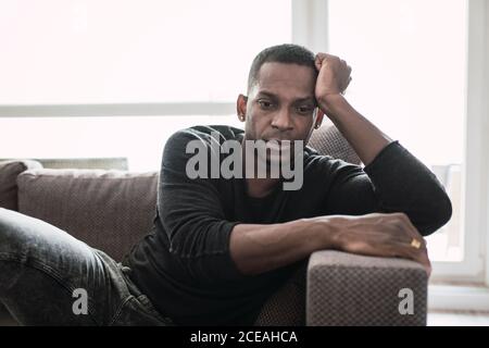 Dreaming adult black man leaning on hands while sitting on couch and looking away in shiny window Stock Photo