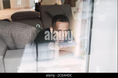 Dreaming adult black man leaning on hands while sitting on couch and looking away in shiny window Stock Photo
